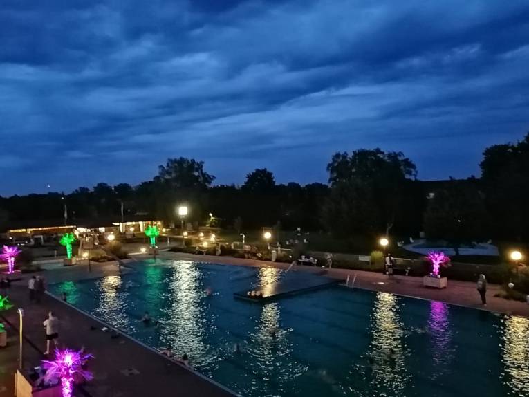 Mit bunten Lichtern und Mondschein beleuchtetes Freibad, Menschen schwimmen oder stehen am Beckenrand